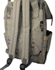 A light beige backpack with a delicate multicolor floral and character print, shown from the back with its padded shoulder straps resting on white bedding.