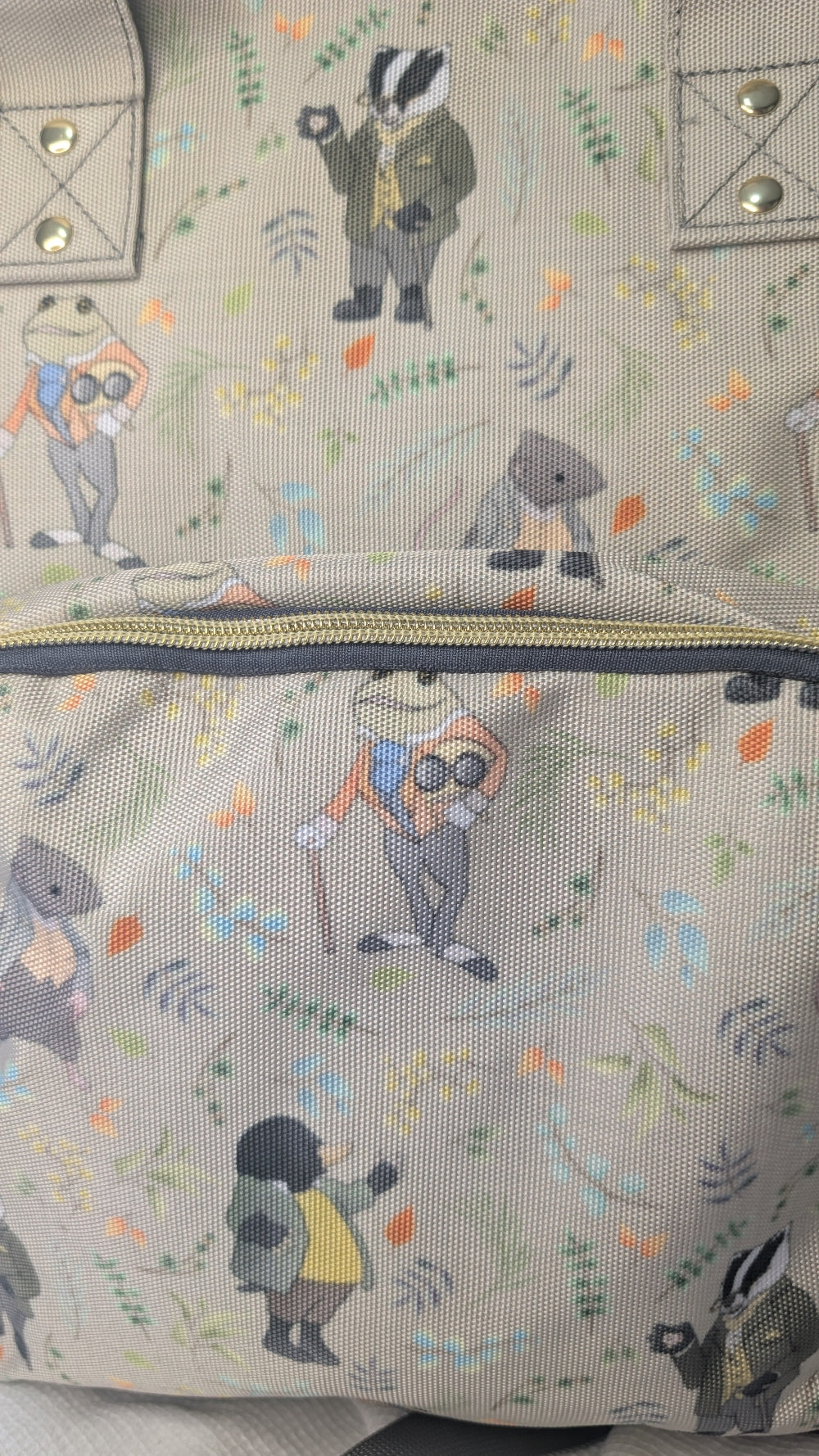 Close-up of a beige canvas tote bag with an all-over print of cartoon animals, including badgers, a frog, and a bird, in muted green, blue, orange, and gray tones.