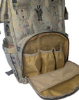 A beige and gray patterned diaper backpack with an open front pocket showing a gold interior organizer, photographed on a white fabric surface.